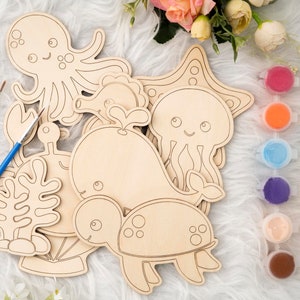May include: A set of wooden sea creature shapes for painting, including an octopus, a whale, a jellyfish, a seahorse, a turtle, and a starfish. The shapes are unpainted and ready to be decorated with paint. There are 10 small jars of paint in various colors.