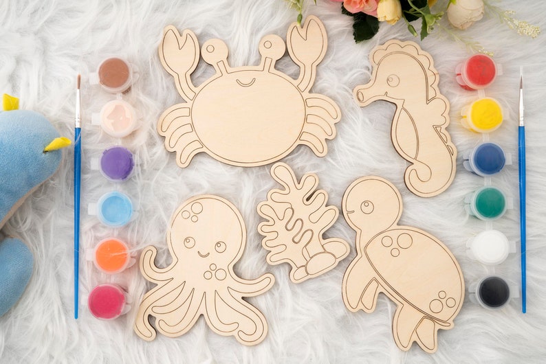 May include: A set of six unfinished wooden sea creature shapes for painting. The set includes a crab, a seahorse, an octopus, a turtle, and two pieces of seaweed. The shapes are all white and have black outlines. There are also ten small jars of paint in various colors.