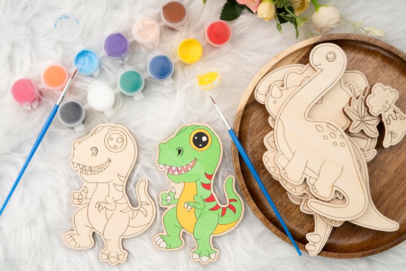 May include: Wooden dinosaur cutouts and paint set. The set includes several unpainted dinosaur shapes, paint pots in various colors, and paintbrushes. One dinosaur is painted green, yellow, and red. The set is designed for arts and crafts.