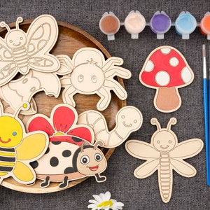 Puede incluir: Un conjunto de formas artesanales de madera con varios insectos, un hongo y una flor. Las formas están sin pintar y listas para ser decoradas con pintura. El conjunto incluye botes de pintura de varios colores y dos pinceles.
