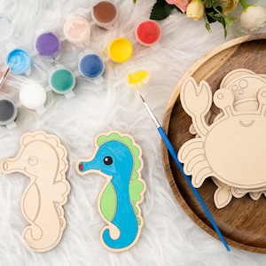 May include: A set of wooden craft kits featuring a seahorse, a crab, and paint pots. The seahorse is light blue with green accents, and the crab is natural wood. The paint pots are various colors, including red, blue, green, yellow, and white.