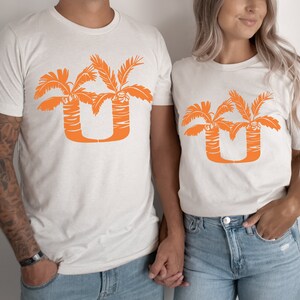 May include: Two white t-shirts with an orange palm tree design that forms the letter "U".
