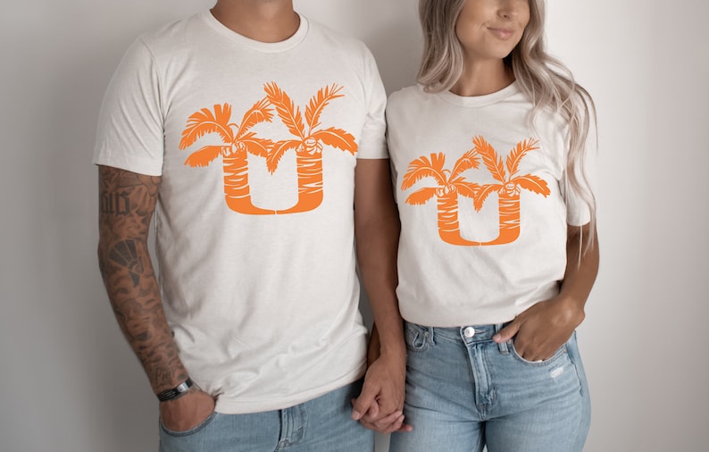 May include: Two white t-shirts with an orange palm tree design that forms the letter "U".