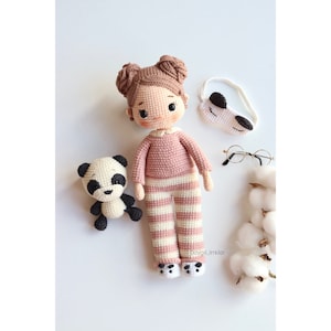 Könnte beinhalten: Eine gehäkelte Puppe, die einen rosa-weiß gestreiften Schlafanzug und eine Schlafmaske trägt. Die Puppe hat braunes Haar und braune Augen. Die Puppe sitzt neben einem gehäkelten Pandabären.