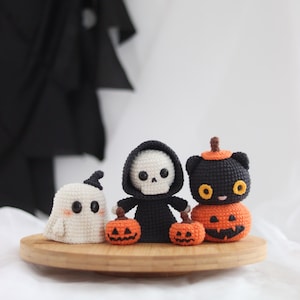 May include: Crocheted Halloween figures including a white ghost, a black Grim Reaper, and a black cat atop a carved pumpkin. The figures are on a wooden tray, perfect for seasonal decor. The Grim Reaper holds two small orange pumpkins.