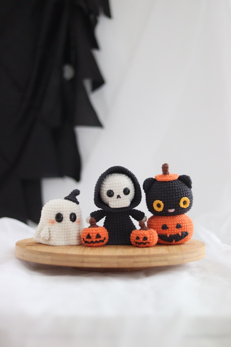 May include: Crocheted Halloween figures including a white ghost, a black Grim Reaper, and a black cat atop a carved pumpkin. The figures are on a wooden tray, perfect for seasonal decor. The Grim Reaper holds two small orange pumpkins.