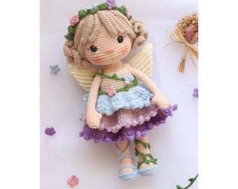 May include: A crocheted fairy doll with blonde hair, a floral crown, and a multi-layered dress with purple, blue, and green accents. The doll has wings and is wearing green vine-like leg warmers.