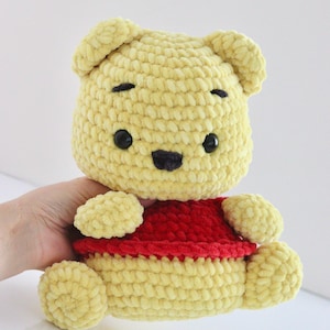 May include: A handmade yellow crochet bear with black eyes and a black nose. The bear is wearing a red shirt. The bear's arms and legs are also yellow. The bear is a plush toy.