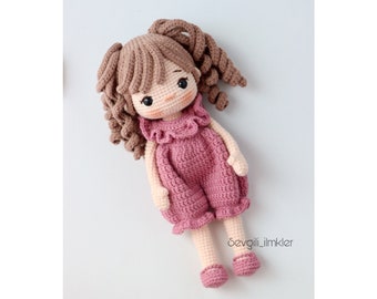 May include: A crocheted doll with brown hair in pigtails wearing a pink dress and pink shoes. The doll is lying on a white surface. The text "Sevgili_imkler" is visible in the bottom right corner of the image.