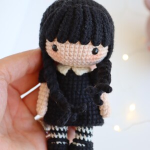 May include: Crocheted doll featuring a black dress, black hair, and white collar. The doll has black and white striped socks and black shoes. The doll is handmade and has a cute, cartoonish appearance.