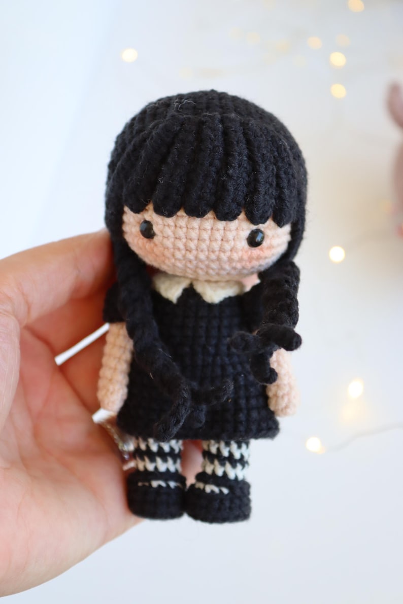 May include: Crocheted doll featuring a black dress, black hair, and white collar. The doll has black and white striped socks and black shoes. The doll is handmade and has a cute, cartoonish appearance.