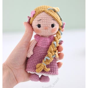 May include: A handmade crochet doll in a pink dress and shoes, with blonde hair in a long braid. The doll has a light skin tone, black eyes, and a small nose. The braid is decorated with colourful accents. The doll is held in a hand.