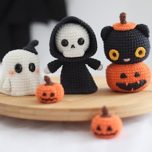 May include: A collection of handmade crochet Halloween decorations. The set includes a white ghost, a grim reaper, a black cat atop a carved pumpkin, and two small pumpkins. The figures are displayed on a wooden tray, perfect for seasonal decor.