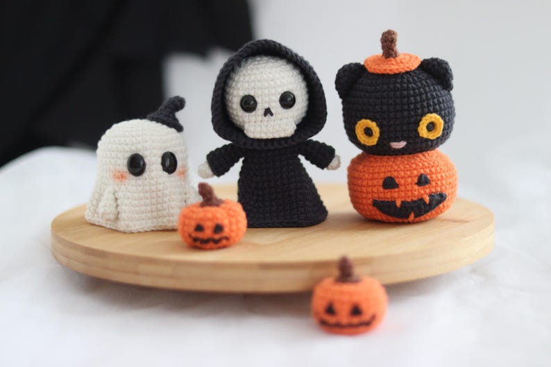 May include: A collection of handmade crochet Halloween decorations. The set includes a white ghost, a grim reaper, a black cat atop a carved pumpkin, and two small pumpkins. The figures are displayed on a wooden tray, perfect for seasonal decor.
