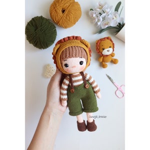 Puede incluir: Una muñeca de crochet con cabello castaño, que lleva un gorro de león marrón y naranja, una camisa a rayas y un mono verde. La muñeca sostiene un pequeño león de crochet.