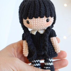 May include: A handmade crochet doll with black hair styled in two braids. The doll wears a black dress with a white collar and black and white striped socks. The doll's skin is a light peach color, and it has black eyes.