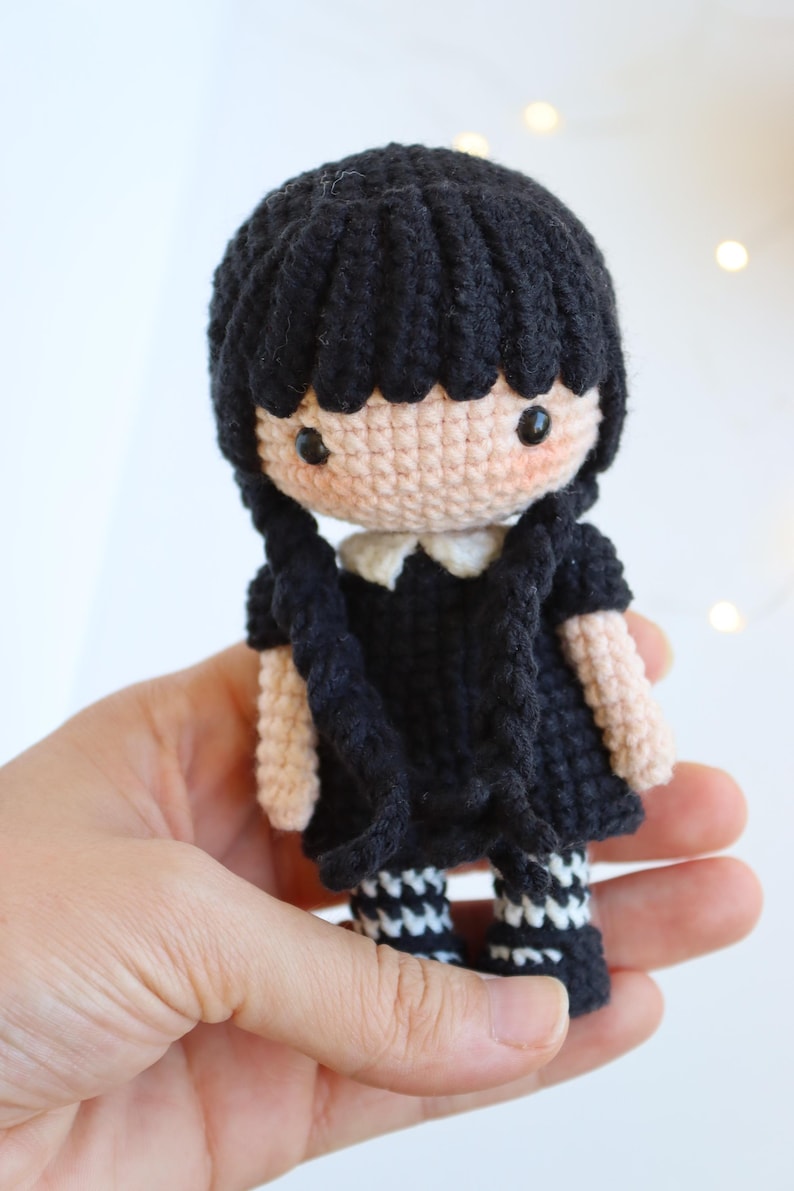 May include: A handmade crochet doll with black hair styled in two braids. The doll wears a black dress with a white collar and black and white striped socks. The doll's skin is a light peach color, and it has black eyes.