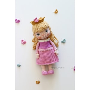 Puede incluir: Una muñeca de crochet con cabello rubio, un vestido rosa y una corona dorada. La muñeca está rodeada de pequeñas mariposas de crochet en varios colores.