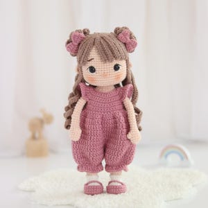 Può includere: Bambola all'uncinetto fatta a mano, con capelli castani e un completo rosa. La bambola indossa una tuta rosa e scarpe abbinate, con due piccoli fiocchi tra i capelli. Ha un'espressione dolce e si trova su una superficie bianca e soffice.