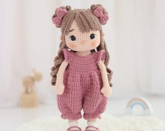 May include: A handmade crochet doll with brown hair and a pink outfit. The doll has a sweet expression and is wearing a pink jumpsuit and matching shoes. The doll's hair is styled with two small bows. The doll is standing on a white, fluffy surface.