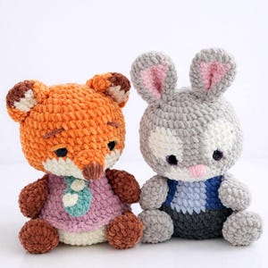 May include: Two handmade crochet stuffed animals: an orange fox wearing a pink dress and a gray bunny with pink inner ears. The fox has brown accents, and the bunny has a blue and gray outfit. Both are sitting.