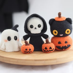 May include: A collection of handmade crochet Halloween decorations, including a ghost, a skeleton in a black robe, a black cat on a pumpkin, and two small jack-o'-lanterns. The figures are displayed on a round wooden tray.