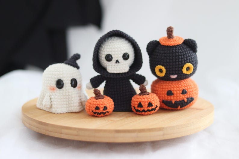 May include: A collection of handmade crochet Halloween decorations, including a ghost, a skeleton in a black robe, a black cat on a pumpkin, and two small jack-o'-lanterns. The figures are displayed on a round wooden tray.