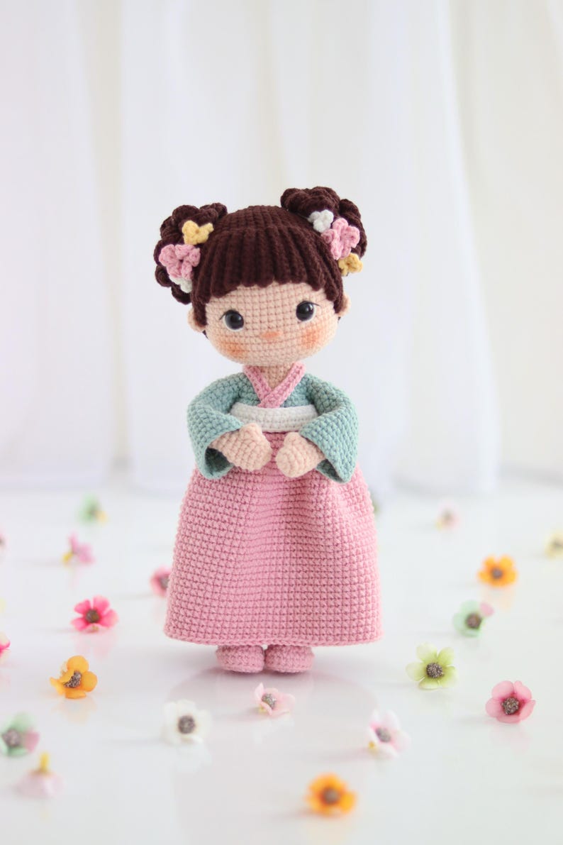 Puede incluir: Una muñeca de ganchillo hecha a mano con un hanbok coreano tradicional. La muñeca tiene cabello castaño peinado en dos moños adornados con flores. El hanbok presenta una parte superior azul claro y una falda rosa. Pequeñas flores están esparcidas alrededor de la muñeca.
