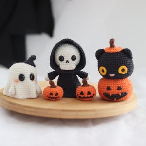 May include: A collection of handmade crochet Halloween decorations. Includes a white ghost, a skeleton in a black robe, and a black cat on top of an orange pumpkin. The decorations are displayed on a round wooden tray.