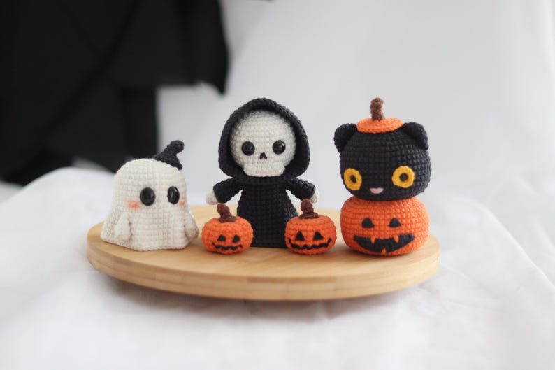 May include: A collection of handmade crochet Halloween decorations. Includes a white ghost, a skeleton in a black robe, and a black cat on top of an orange pumpkin. The decorations are displayed on a round wooden tray.