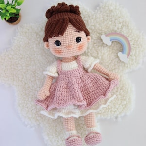 May include: A handmade crochet doll with brown yarn hair styled in a bun, wearing a cream-colored short-sleeved top and a dusty pink pinafore dress. The doll has black button eyes, rosy cheeks, and matching pink shoes with white socks.