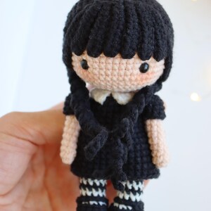 May include: Crocheted doll with black hair styled in two braids. The doll wears a black dress with a white collar and black and white striped socks. The doll's skin is a light peach color, and it has rosy cheeks.