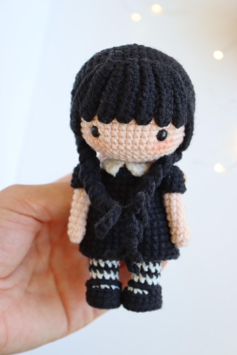 May include: Crocheted doll with black hair styled in two braids. The doll wears a black dress with a white collar and black and white striped socks. The doll's skin is a light peach color, and it has rosy cheeks.