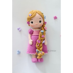 May include: A crocheted doll with long blonde hair braided with flowers. The doll is wearing a pink dress and has a sweet expression on its face.