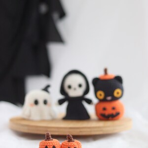 May include: Crocheted Halloween-themed decorations. Includes a white ghost, a black grim reaper, and a black cat atop an orange pumpkin. Two small orange pumpkins with carved faces are in the foreground. All are displayed on a wooden tray.