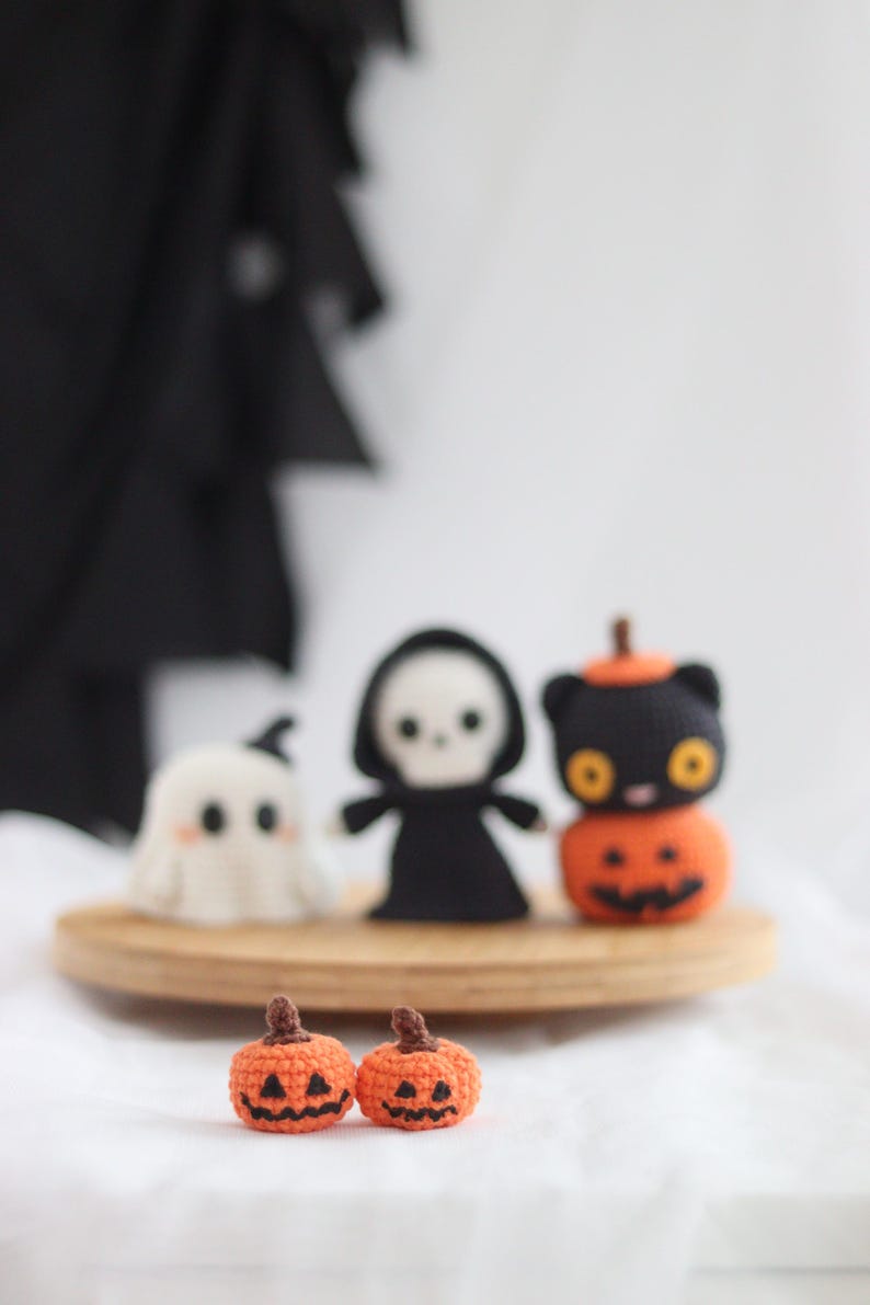 May include: Crocheted Halloween-themed decorations. Includes a white ghost, a black grim reaper, and a black cat atop an orange pumpkin. Two small orange pumpkins with carved faces are in the foreground. All are displayed on a wooden tray.