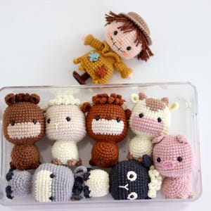 May include: A collection of handmade crochet farm animal toys, including a horse, sheep, cow, pig, and a scarecrow doll. The toys are in a clear plastic container. The scarecrow doll is wearing a yellow outfit and a brown hat.