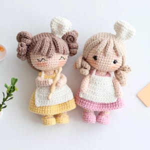 May include: Two crocheted dolls wearing aprons and chef hats. One doll is wearing a yellow dress and the other is wearing a pink dress.