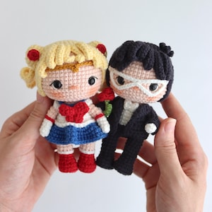 May include: Crocheted dolls of a blonde girl in a sailor outfit with a red heart and a dark-haired man in a suit, holding a red rose. The dolls have black eyes and are handmade.