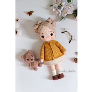 May include: A crocheted doll with blonde hair, wearing a yellow cardigan and a white skirt, stands with a brown crocheted teddy bear. The doll has brown eyes and rosy cheeks. The background is white with a few decorative items.