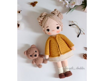 May include: A crocheted doll with blonde hair, wearing a yellow cardigan and a white skirt, stands with a brown crocheted teddy bear. The doll has brown eyes and rosy cheeks. The background is white with a few decorative items.