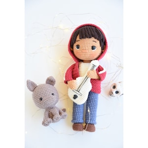 May include: A crocheted doll with brown hair, wearing a red hoodie, blue jeans, and holding a white guitar. A small crocheted pig sits in front of the doll.