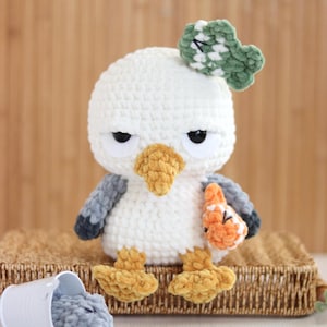 May include: A white and gray crocheted seagull plush toy with a yellow beak and feet. The seagull has a green crocheted fish on its head and holds an orange fish in its wing. A small white bucket containing gray yarn pieces sits nearby.