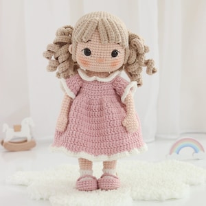 Puede incluir: Muñeca de ganchillo hecha a mano con un vestido rosa, cuello blanco y zapatos a juego. La muñeca tiene coletas rubias y está de pie sobre una superficie blanca y esponjosa. Un juguete encantador y artesanal.
