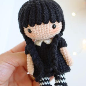May include: A handmade crochet doll with black hair styled in two braids. The doll wears a black dress with a white collar and black and white striped socks. The doll's skin is a light peach color, and it has rosy cheeks.