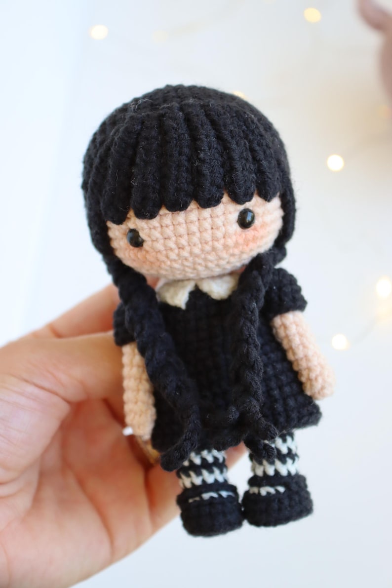 May include: A handmade crochet doll with black hair styled in two braids. The doll wears a black dress with a white collar and black and white striped socks. The doll's skin is a light peach color, and it has rosy cheeks.