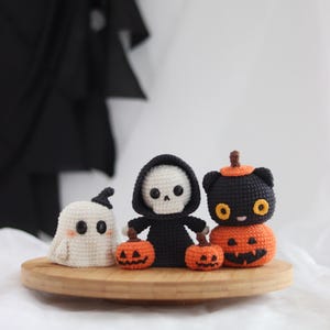 May include: Crocheted Halloween decorations on a wooden tray. Includes a white ghost, a black-robed skeleton holding two orange pumpkins, and a black cat atop an orange pumpkin. The decorations are handmade and suitable for Halloween.