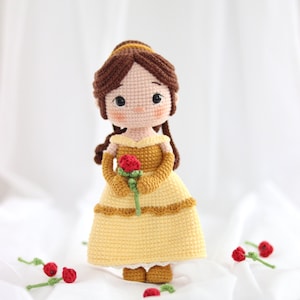 Könnte beinhalten: Handgefertigte Häkelpuppe einer Prinzessin in einem gelben Kleid, die eine rote Rose hält. Die Puppe hat braune Haare und steht auf einer weißen Oberfläche. Mehrere kleine rote Rosen sind um die Puppe herum verstreut.