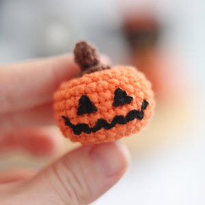 May include: A small, crocheted orange pumpkin with a brown stem and a carved black face. The handmade Halloween decoration is held in a person's hand. The pumpkin is a festive, seasonal item.