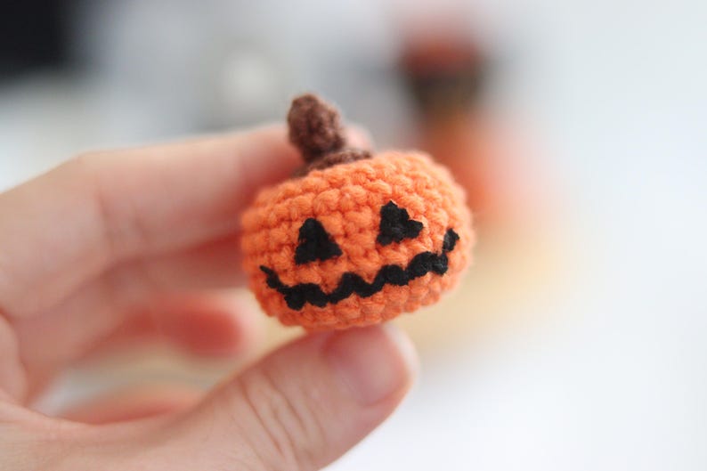 May include: A small, crocheted orange pumpkin with a brown stem and a carved black face. The handmade Halloween decoration is held in a person's hand. The pumpkin is a festive, seasonal item.